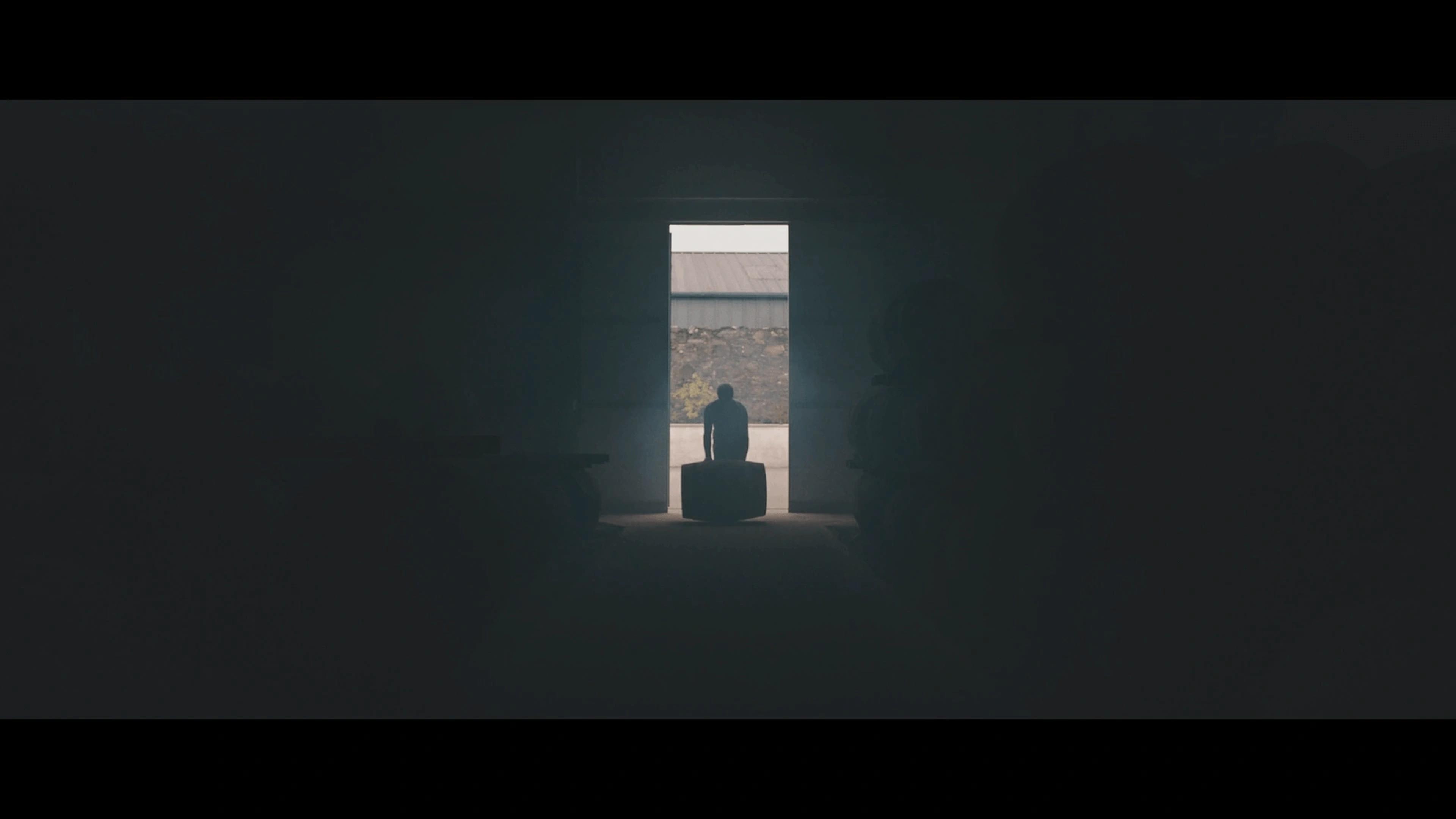 A scene from the 'KILCHOMAN: 100% ISLAY FROM BARLEY TO BOTTLE' film, with cinematography by Paolo Bischi. The image shows the silhouette of a man illuminated by light through an open door as he rolls a whiskey barrel into the distillery.