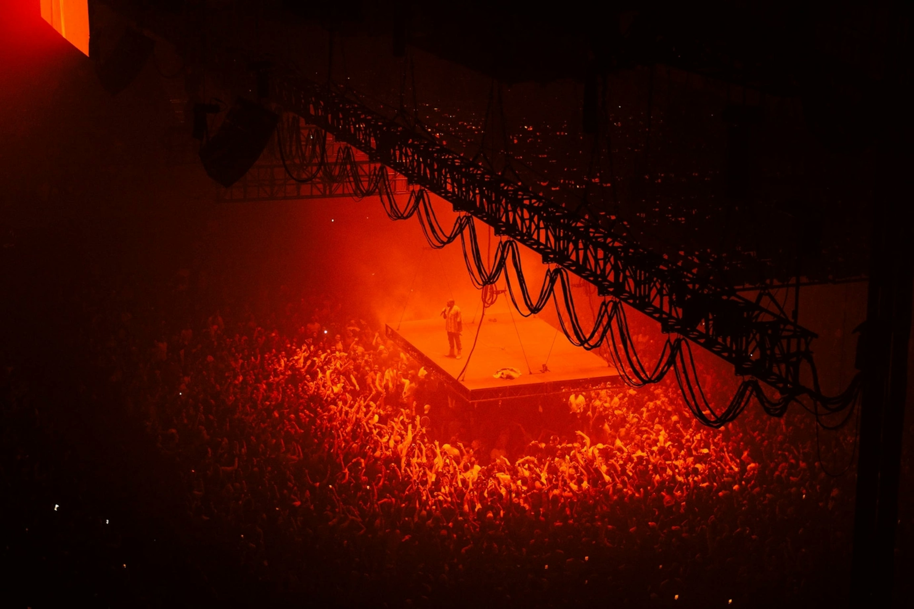 Kanye West performing on stage surrounded by enthusiastic fans, illuminated by vibrant neon red lighting. Image by Karina Carvalho on Unsplash.